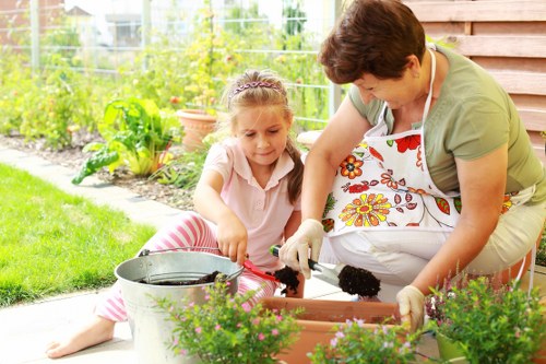 A woman and a young girl are working together in a well-maintained garden, planting or tending to potted plants on a patio area. The woman is wearing a floral apron and gardening gloves, focusing on the task, while the girl, dressed in a light pink top with striped pants, assists by handling a small gardening tool. The garden features lush green grass and a vegetable patch or flower beds with a variety of plants and emerging foliage, bordered by a neat garden edge. In the background, there's a wooden shed with a natural finish, and beyond that, a wire fence encloses the garden space. The scene is set in daylight, with soft natural light illuminating the outdoor environment, implying good weather conditions. The patio surface is made of concrete or stone, and several potted plants with vibrant green foliage and flowering plants are arranged along the edge. The overall scene exudes a peaceful and productive outdoor setting typical of residential garden maintenance, aligned with gardening services in the Chigwell area, as part of a professional landscaping or gardening project.