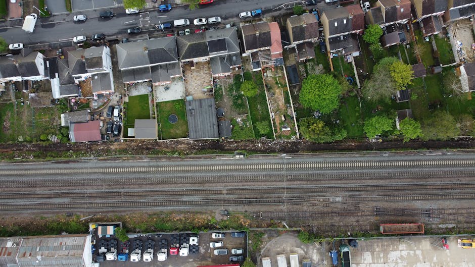 An aerial view of a row of residential gardens along a railway line, displaying various landscaped and maintained outdoor spaces. The gardens feature lush green lawns, some with trimmed grass and flow