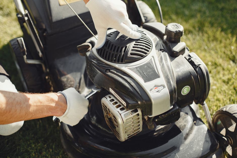 Close-up view of an orange petrol-powered lawn mower resting on a well-maintained grassy garden area. The mower features a black engine cover on top, with visible scratches and some dirt on the machin
